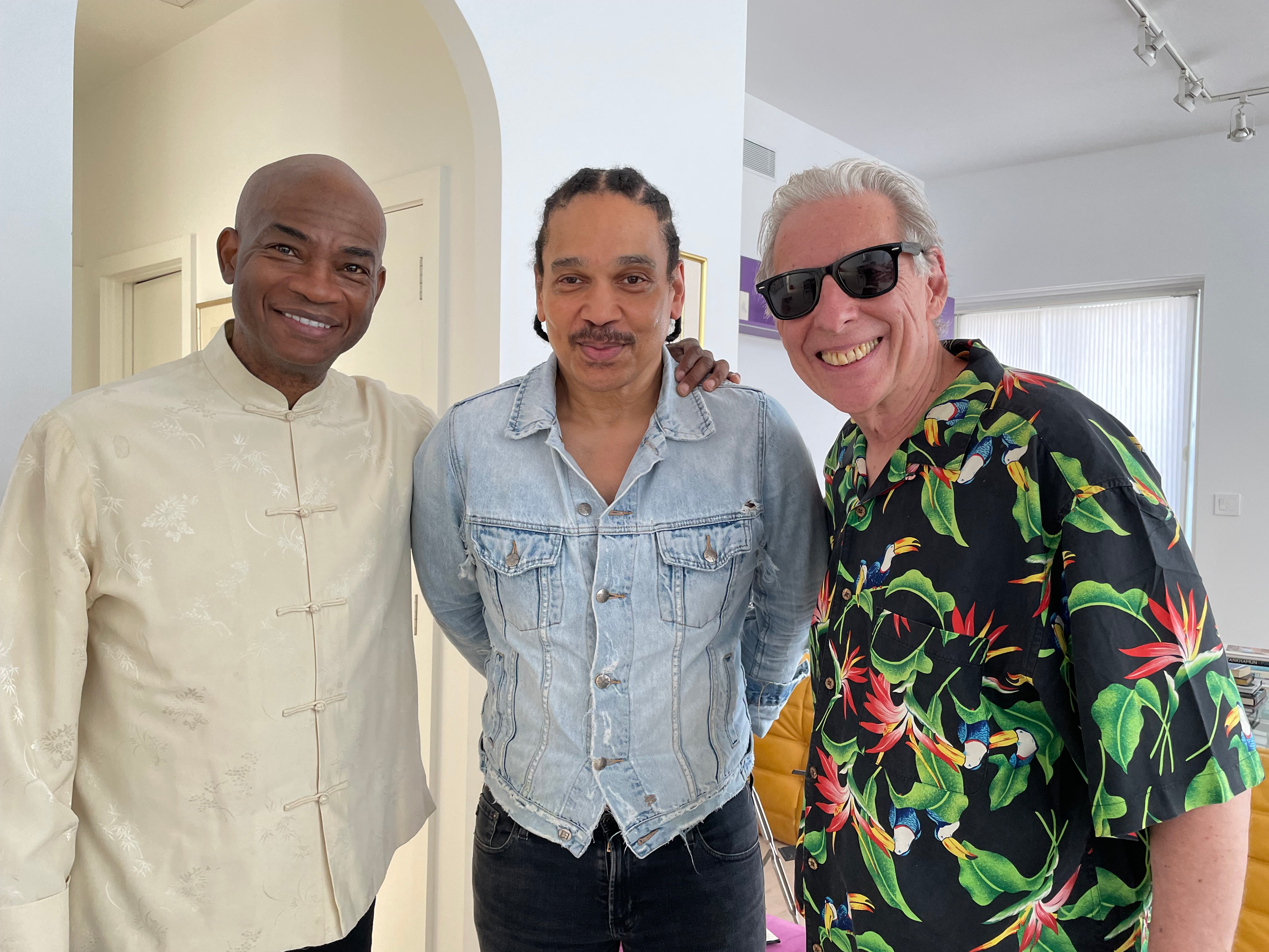 Three men standing together indoors, smiling, with one wearing a cream traditional Asian shirt, one in a light denim jacket, and another in a black tropical shirt with colorful parrots and leaves, with sunglasses.