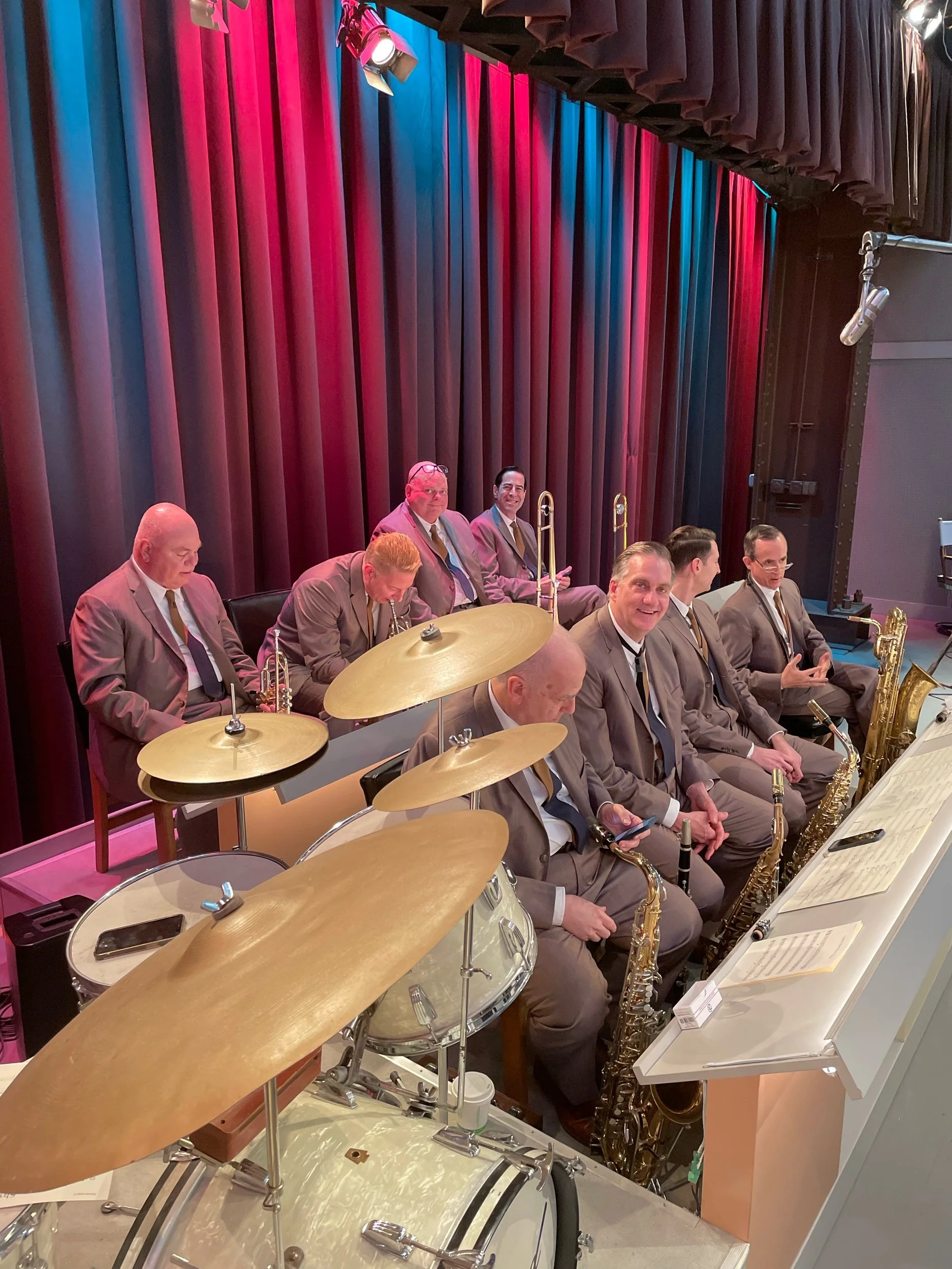 Jazz band in gray suits with accordions, saxophones, and trombones, sitting behind a drum set and music stands, on stage with red and blue curtains.
