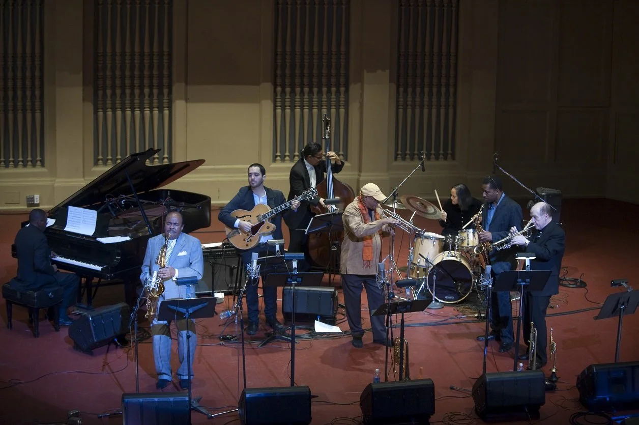 Jazz band performing on stage with piano, saxophone, guitar, bass, drums, and trombones.