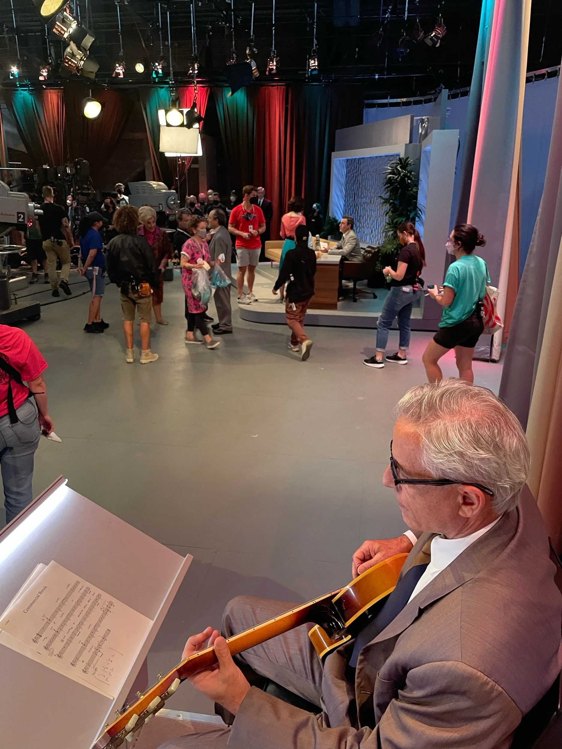 An older man playing guitar with sheet music in front of him, while people are walking and talking in a television studio set.