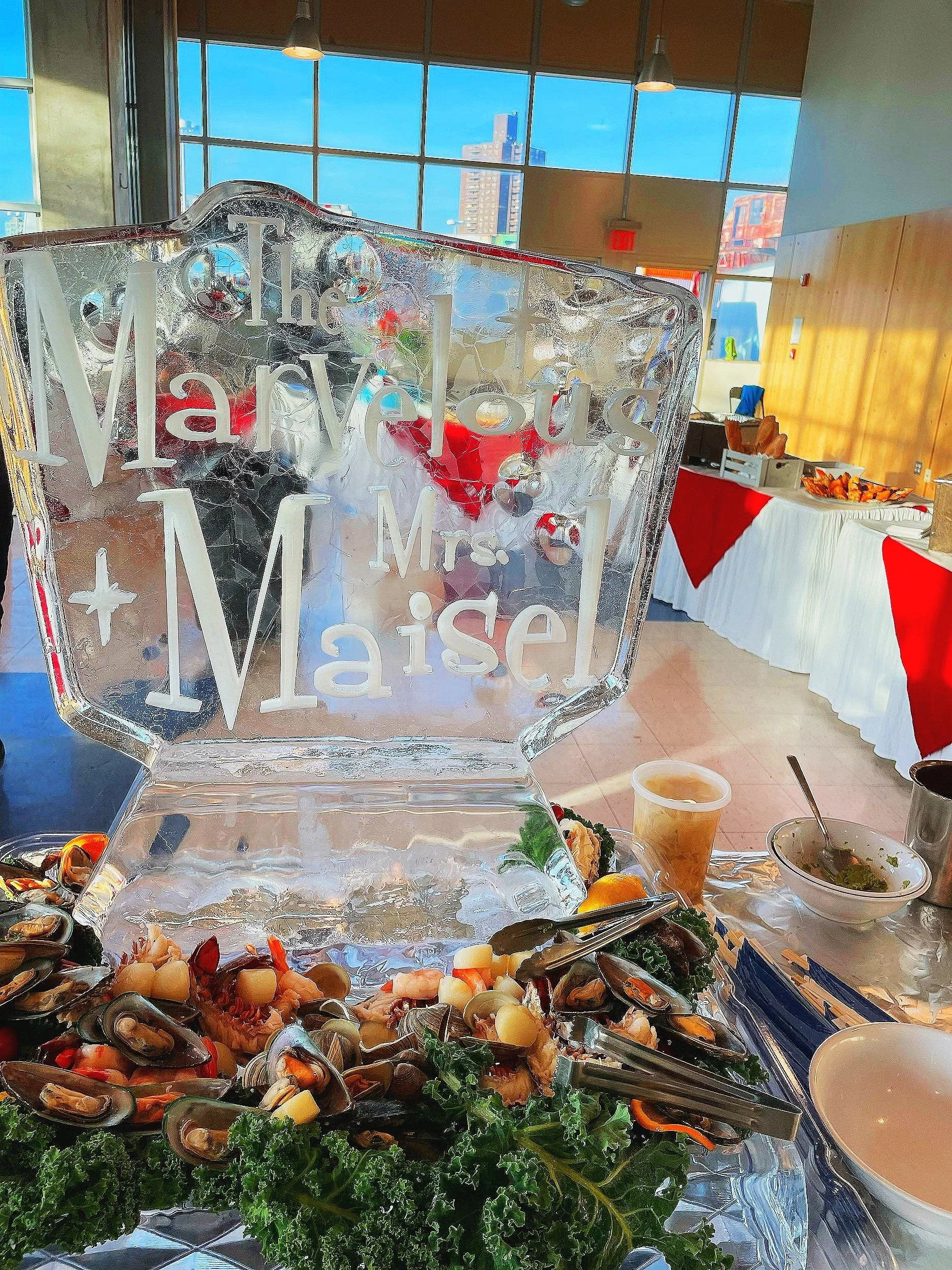 Ice sculpture with engraved words 'The Marvelous Mrs. Maisel' at a restaurant or banquet event, with seafood and salad on the table, and a buffet table in the background.
