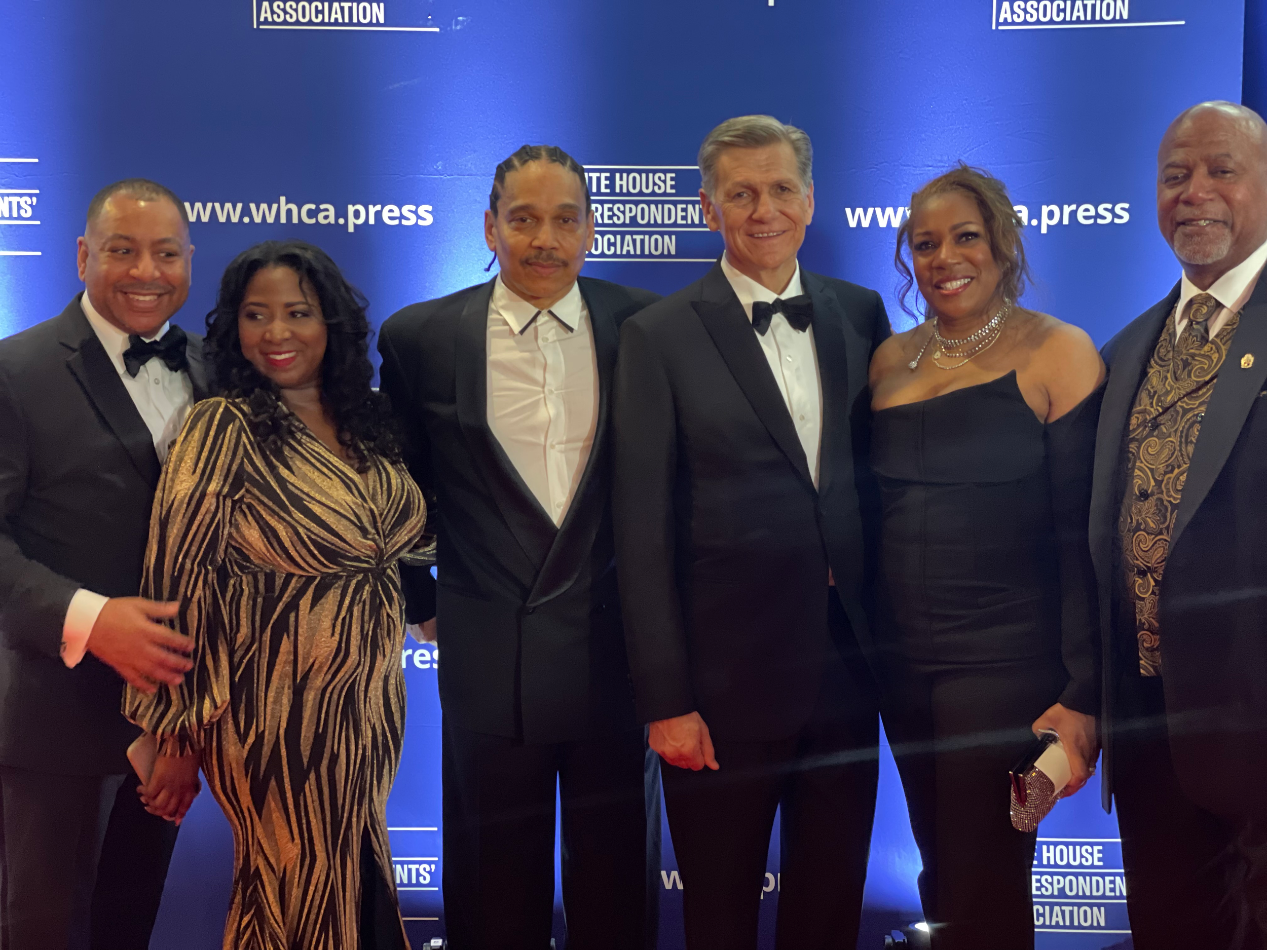 Group of six people dressed in formal evening wear, standing in front of a blue backdrop with white text, at a White House Correspondents' Association event.