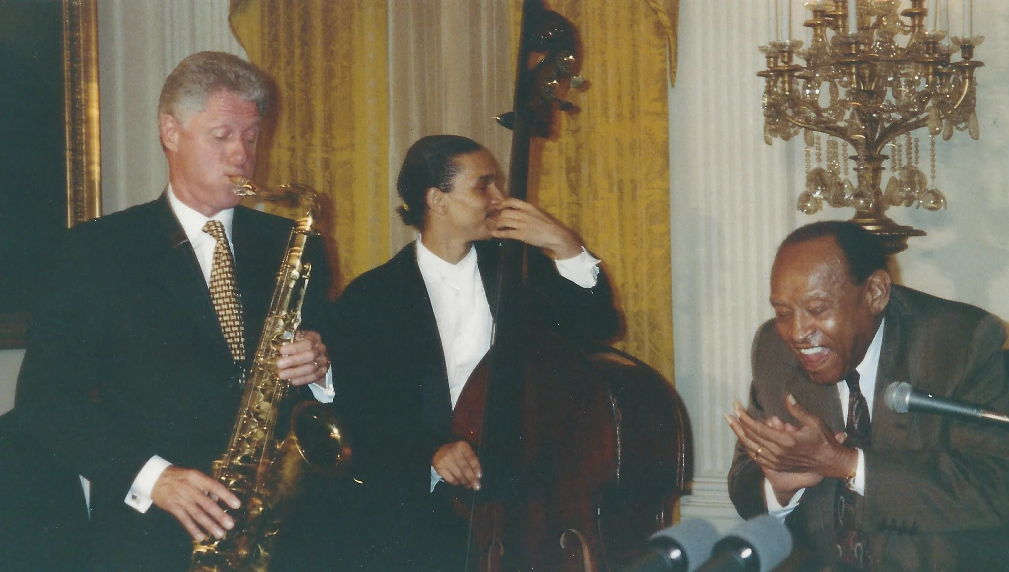 Three people in a formal setting, two men and one woman, with musical instruments and a microphone, engaging in a lively conversation or performance in a room with elegant decor, including a large candelabra and yellow curtains.