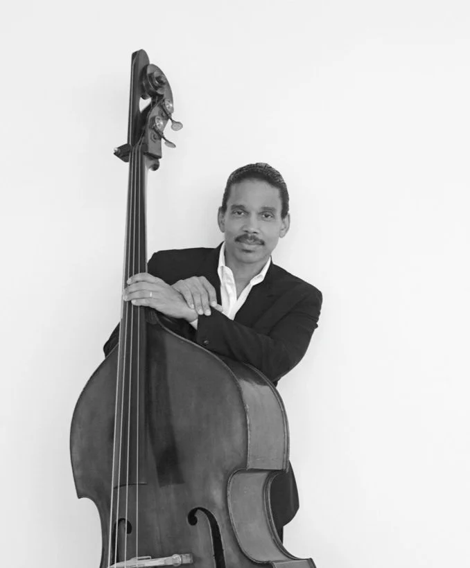 A man in a black suit holding an upright bass in front of a plain white wall.